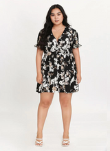 Load image into Gallery viewer, Full-body front view of a black, white, and cream floral mini dress with ruffles at the sleeves styled with white strappy heels.
