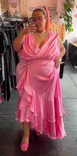Load image into Gallery viewer, Full-body front view of a size 4 Fashion to Figure x Garner Style collab bubblegum pink maxi wrap dress with big ruffle tiers and a wicker-style buckle belt styled without its belt on a size 22/24 model.
